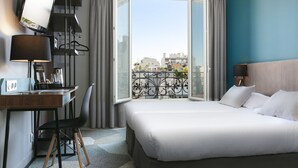 Habitación de hotel con cama, escritorio con computadora, silla, televisor montado en la pared y balcón con vista a edificios.
