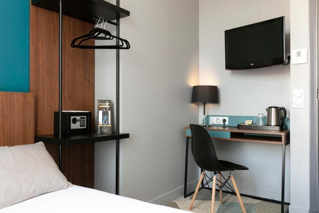 A hotel room with a bed, a desk with a chair, a TV, a lamp, a coffee maker, and a radio.