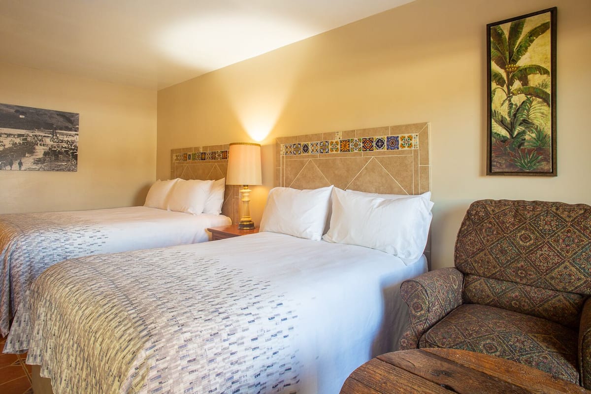 A hotel room with two beds, a nightstand with a lamp, a patterned armchair, and a decorative wall panel.