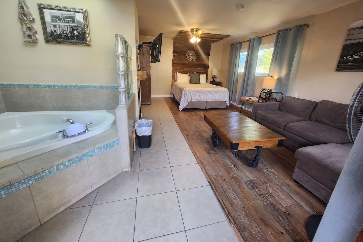 A hotel room with a large bathtub, a wooden floor, a bed with a white bedspread, a brown sofa, and a ceiling fan.