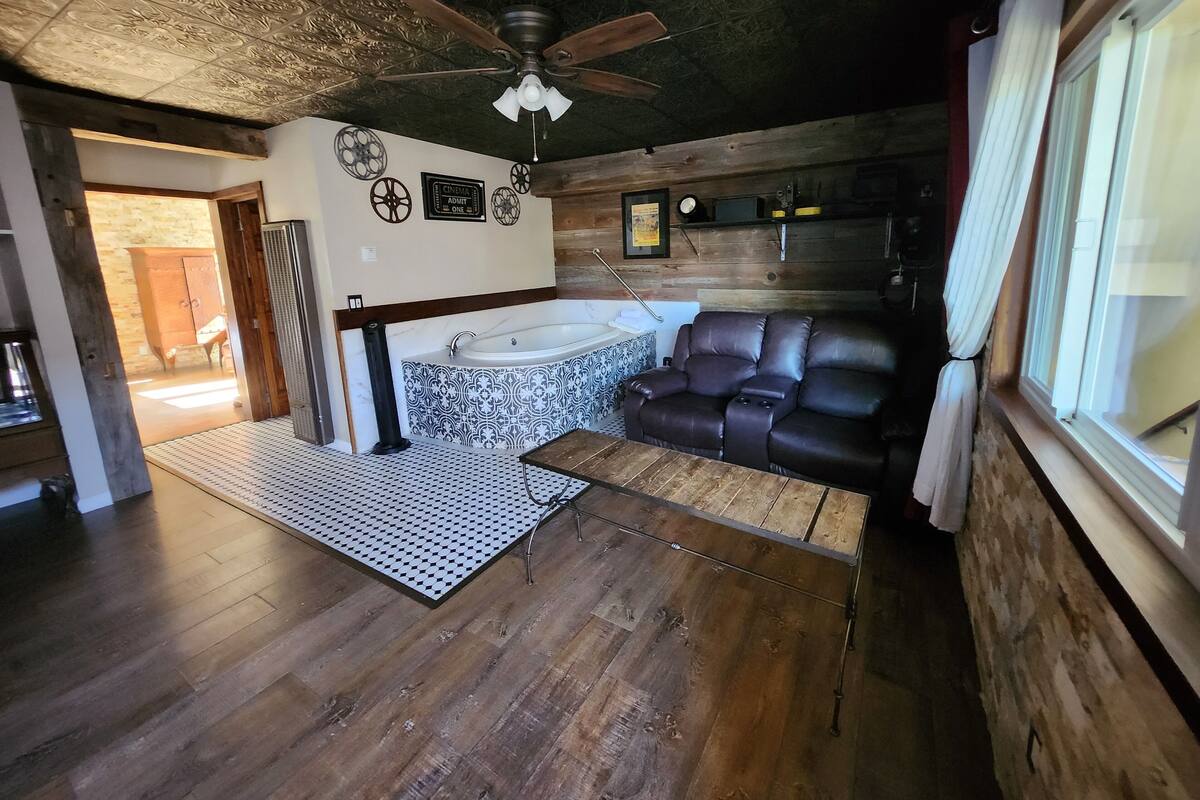 A spacious room with a wooden floor, a ceiling fan, a large window with curtains, a black leather sofa, and a bathtub.