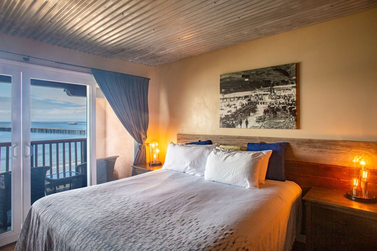 A hotel room with a bed, a nightstand with a lamp, a window with curtains, and a view of the sea.