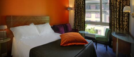 Habitación de hotel con una cama grande, un escritorio, una silla y vistas a edificios desde la ventana.