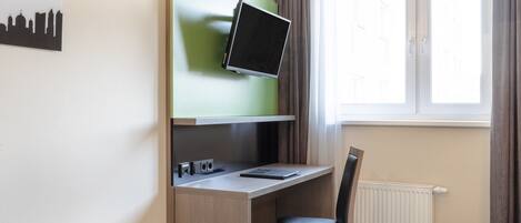 Habitación de hotel con una cama, un escritorio con computadora, una silla, una ventana con cortinas y un televisor instalado en la pared.