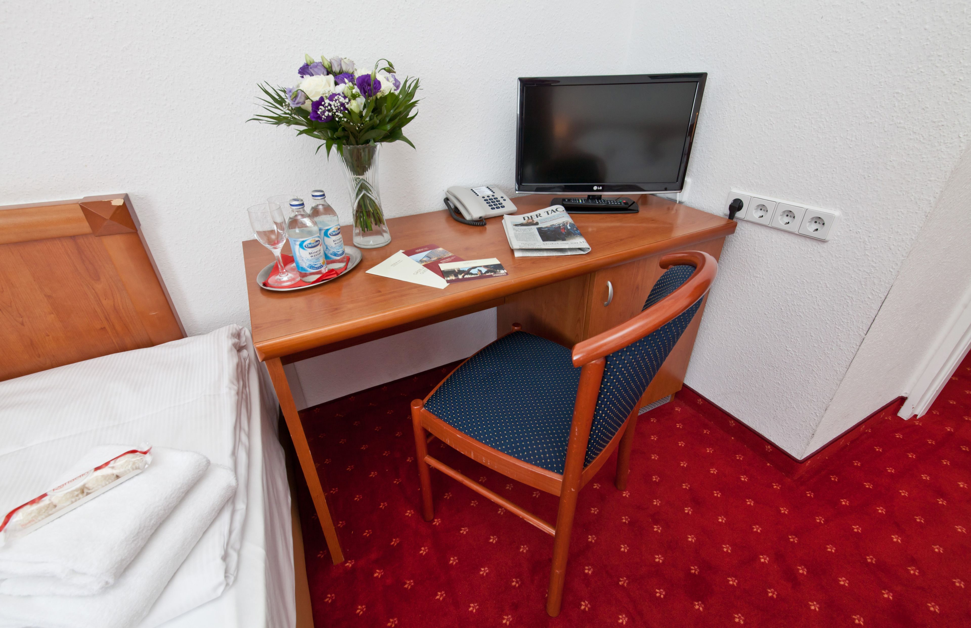 Une chambre d’hôtel avec un lit, un bureau avec un ordinateur, une chaise, un vase avec des fleurs, un téléphone et un journal.