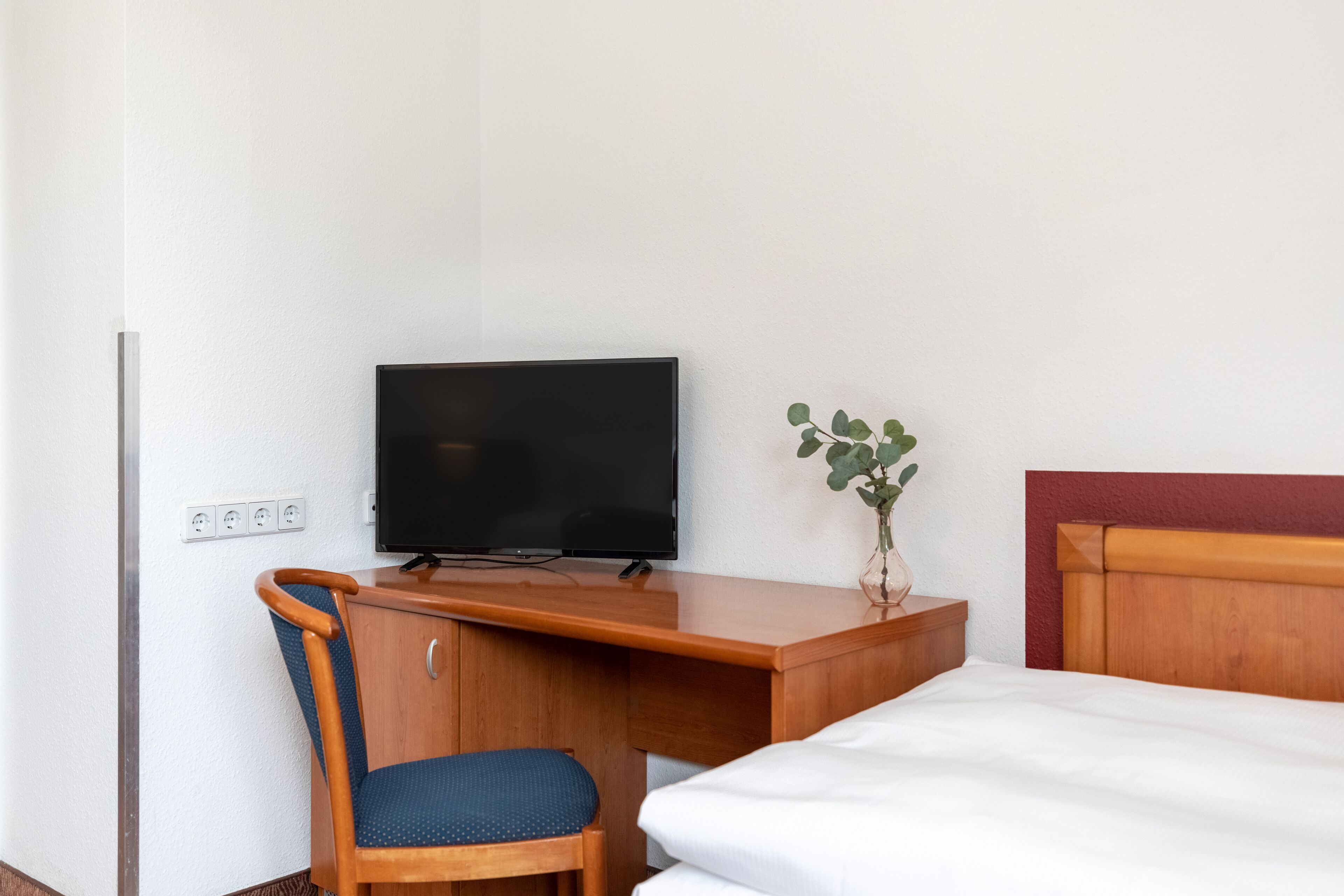 Hotellihuone, jossa on puinen työpöytä, televisio, tuoli, sänky ja pieni kasvi.