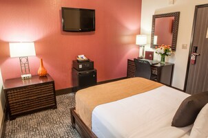 Habitación de hotel con cama, televisor montado en la pared, escritorio con lámpara, jarrón con flores y puerta con manija.