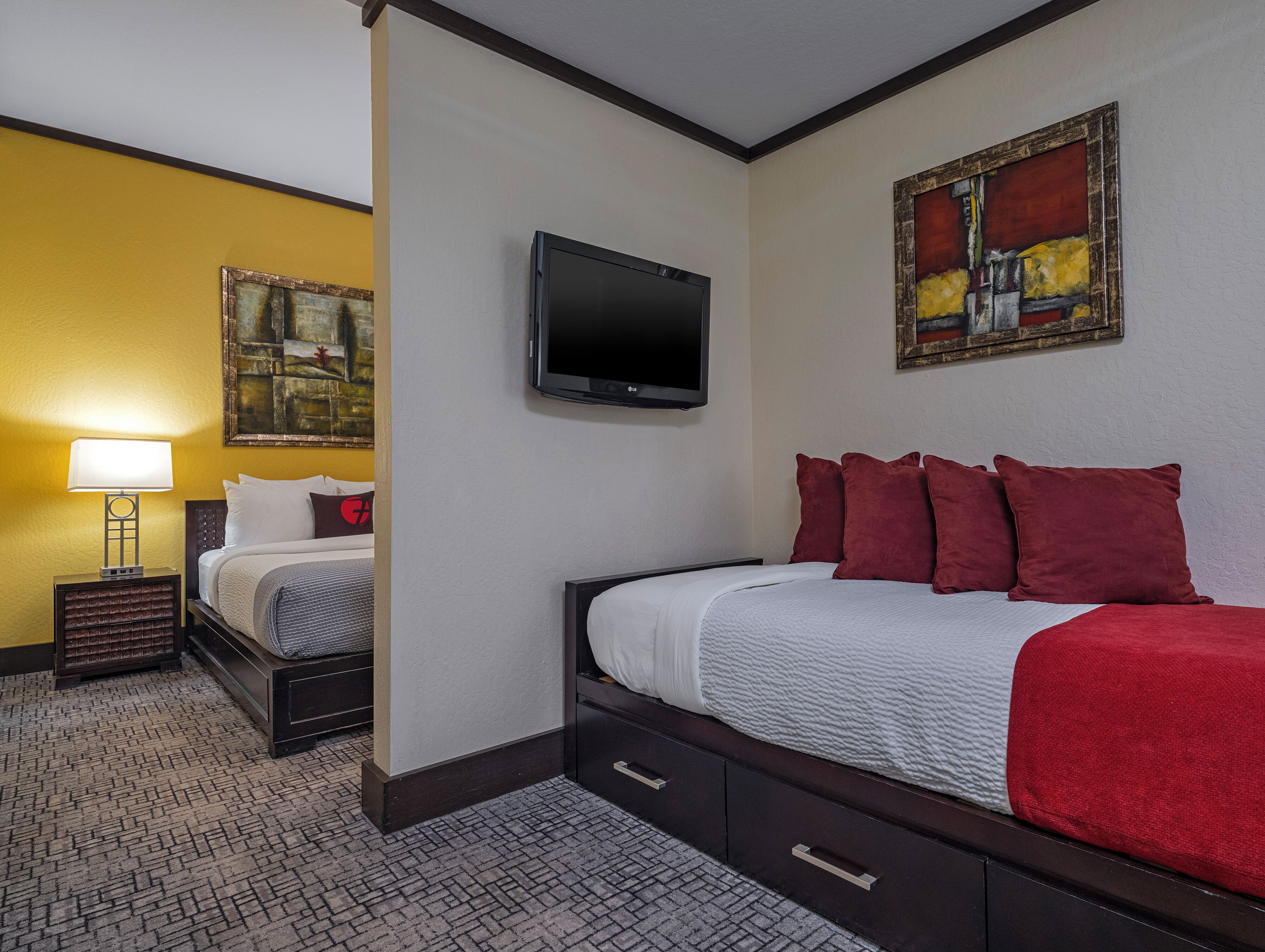 Ein Hotelzimmer mit zwei Betten, einem Fernseher, Kunstwerken an den Wänden und einer Lampe.