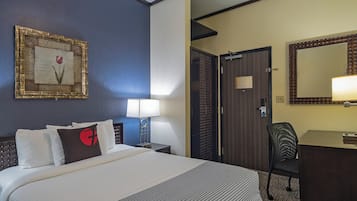 A hotel room with a bed, a desk, a chair, a lamp, a mirror, and a door.