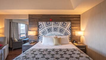 Quarto de hotel com uma cama grande, cabeceira de madeira, duas mesas de cabeceira com abajures, um sofá e vista para a cidade pela janela.