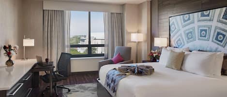 A hotel room with a large bed, a desk with a laptop, a chair, and a view of the city through the window.