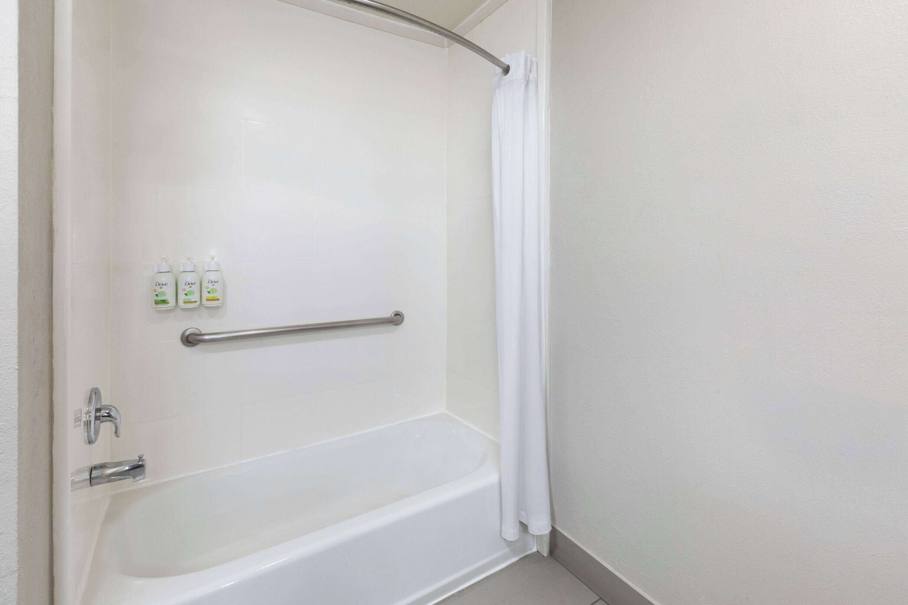 Una bañera blanca con cortina de ducha, una barra de apoyo y dos botellas de Dove para el cuerpo en la pared.