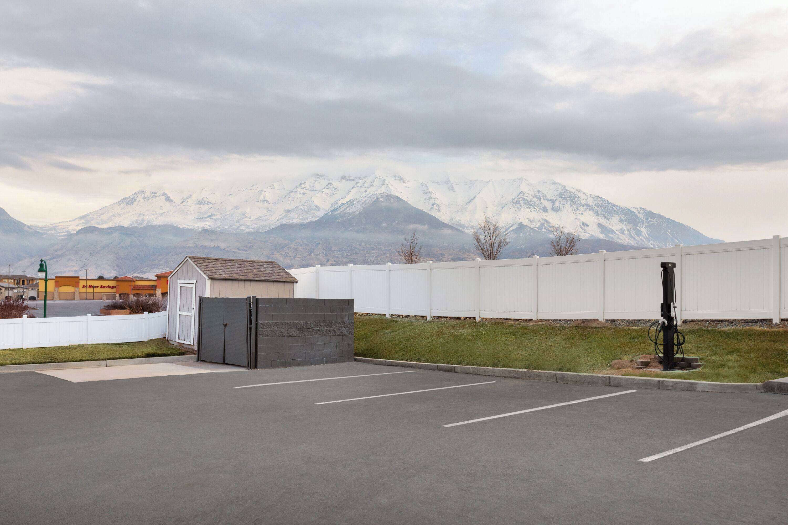 Un estacionamiento con un pequeño edificio, una cerca blanca y una cadena montañosa al fondo.