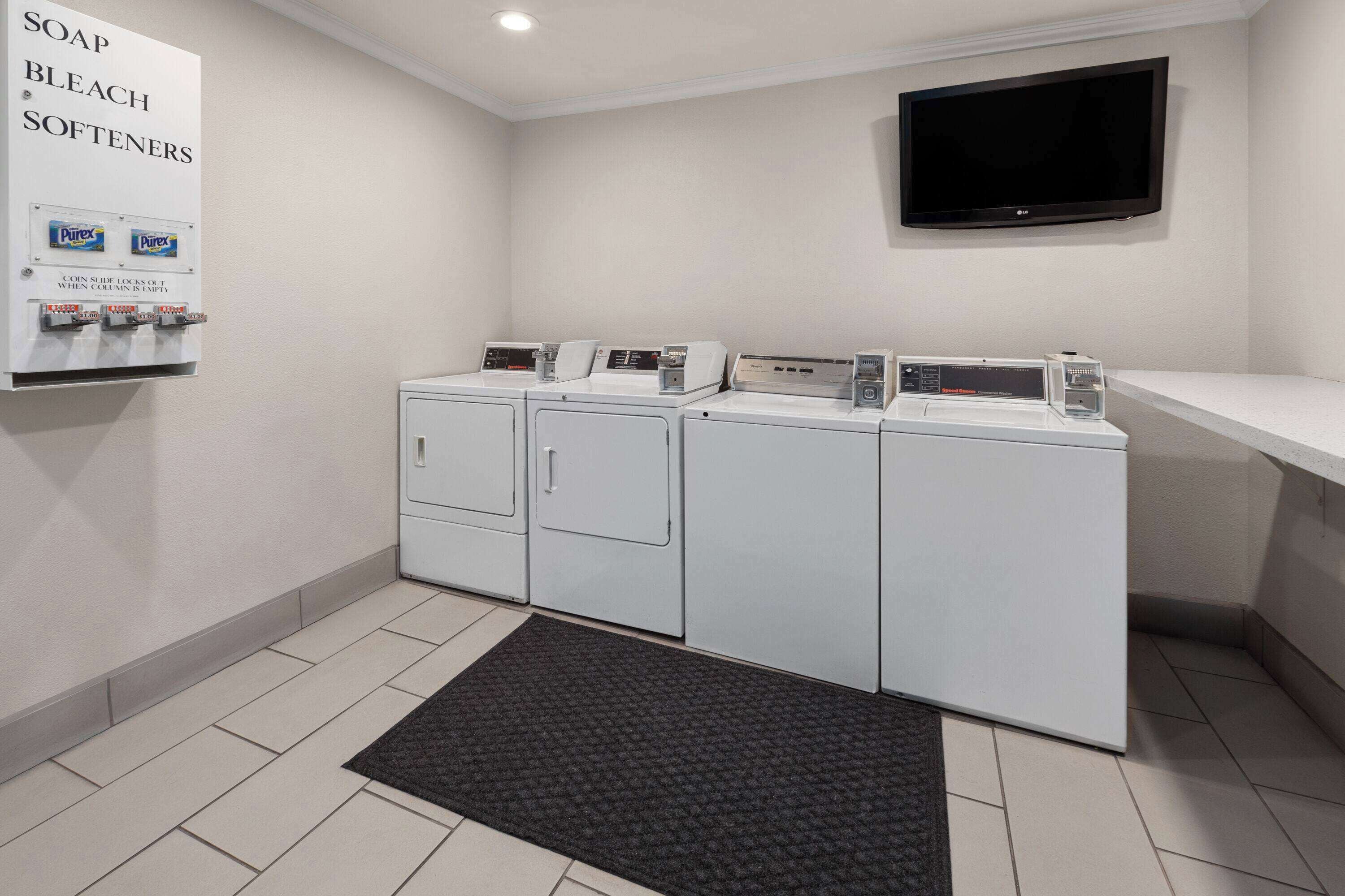 A laundry room with four washing machines, a television mounted on the wall, and a sign listing available laundry supplies.
