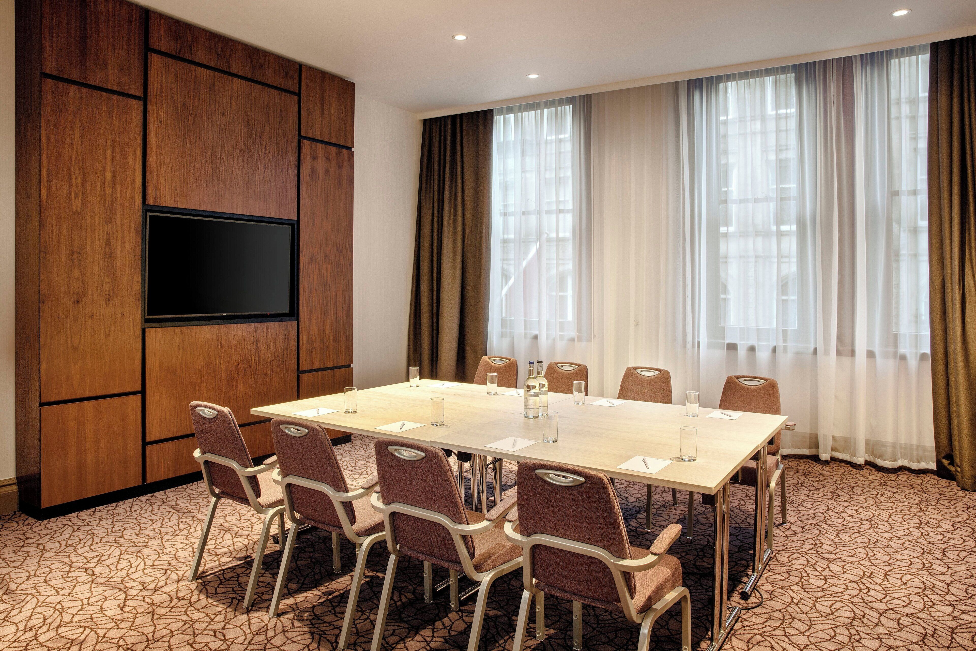 A conference room with a long table, chairs, a flat-screen TV, and large windows with curtains.