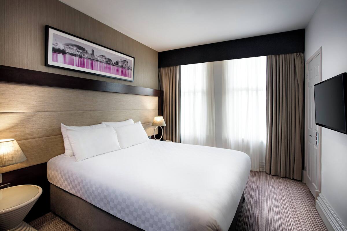 A hotel room with a large bed, a bedside table with a lamp, a flat-screen TV, and a framed picture on the wall.