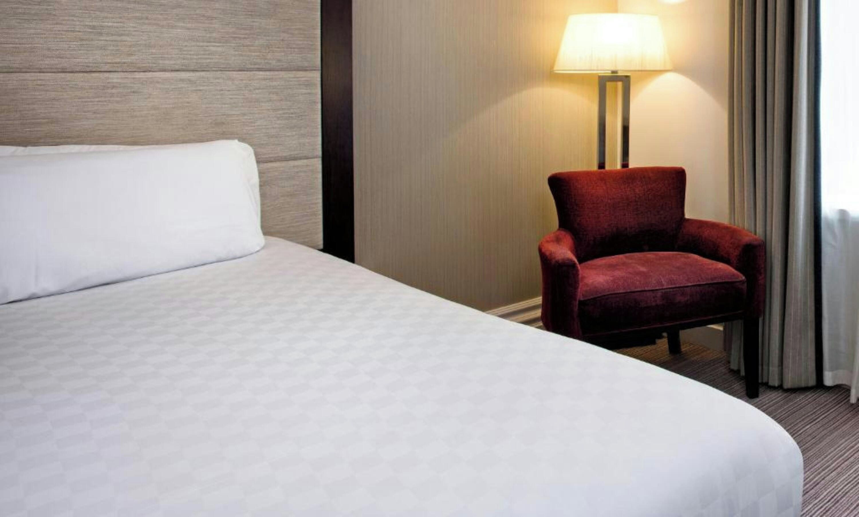 Une chambre d’hôtel avec un lit, un fauteuil rouge, une lampe et une fenêtre avec des rideaux.