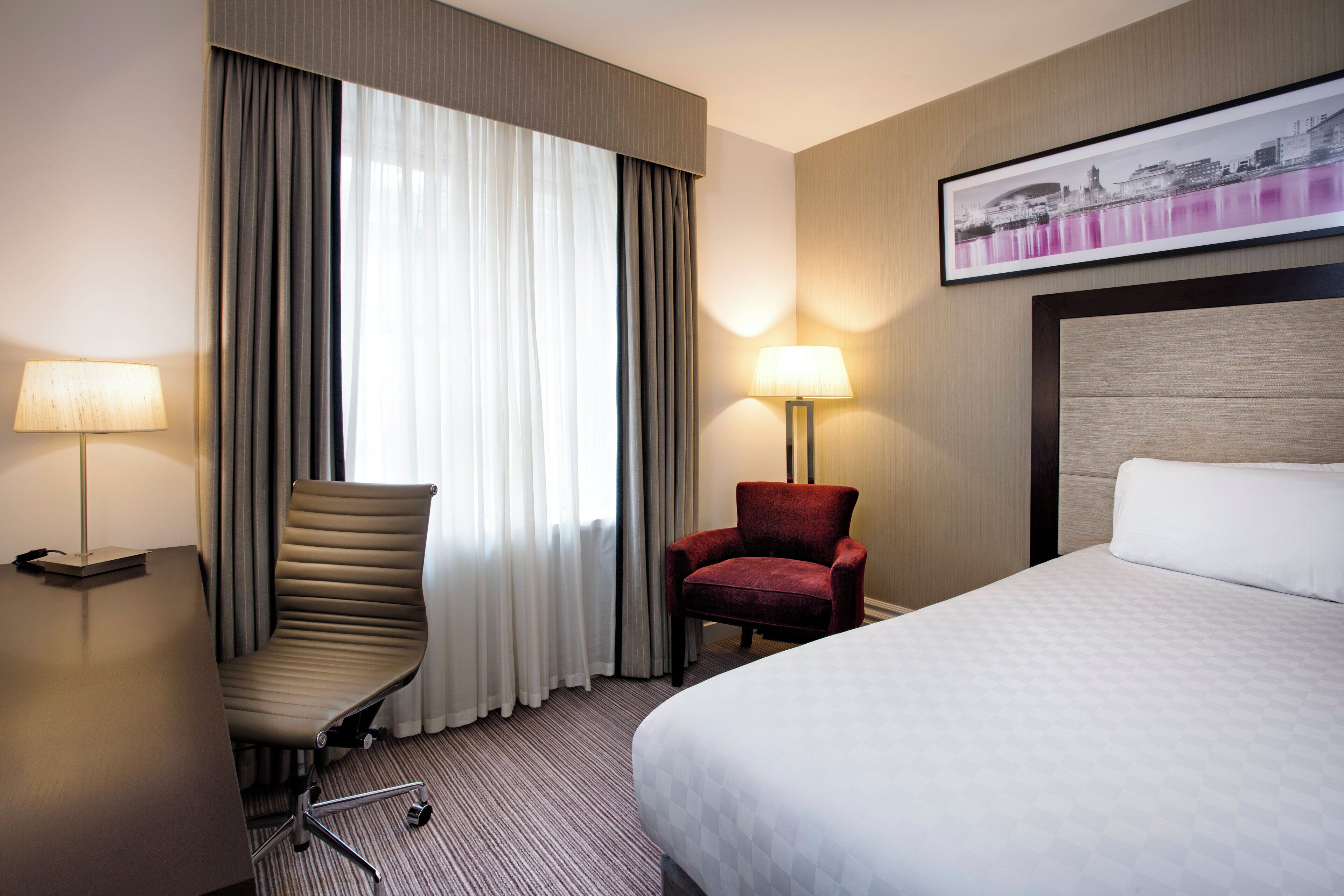 Une chambre d’hôtel avec un lit, deux chaises, un bureau avec une lampe, une grande fenêtre avec des rideaux et un tableau encadré au mur.