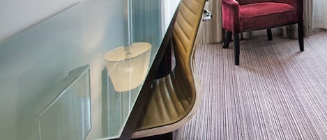 A hotel room with a glass desk, a red chair, a lamp, and a flat-screen TV mounted on the wall.