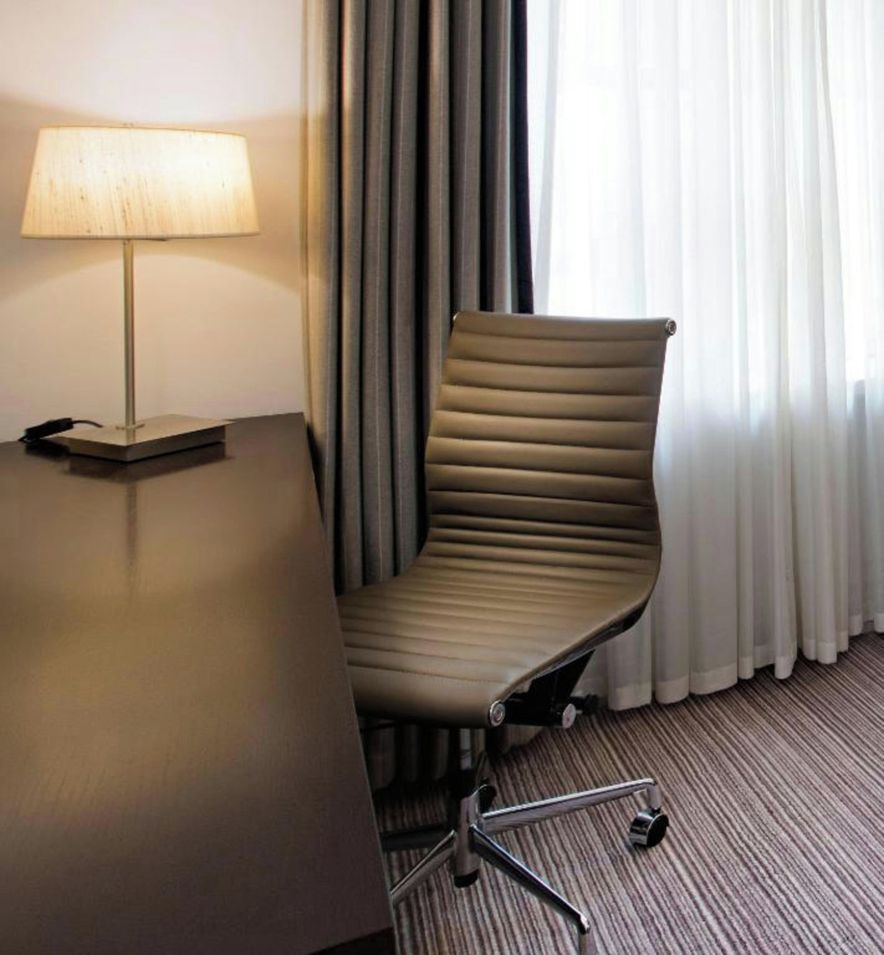Un fauteuil de bureau moderne, doté d’un haut dossier et d’accoudoirs, placé à côté d’un bureau sur lequel se trouve une lampe.