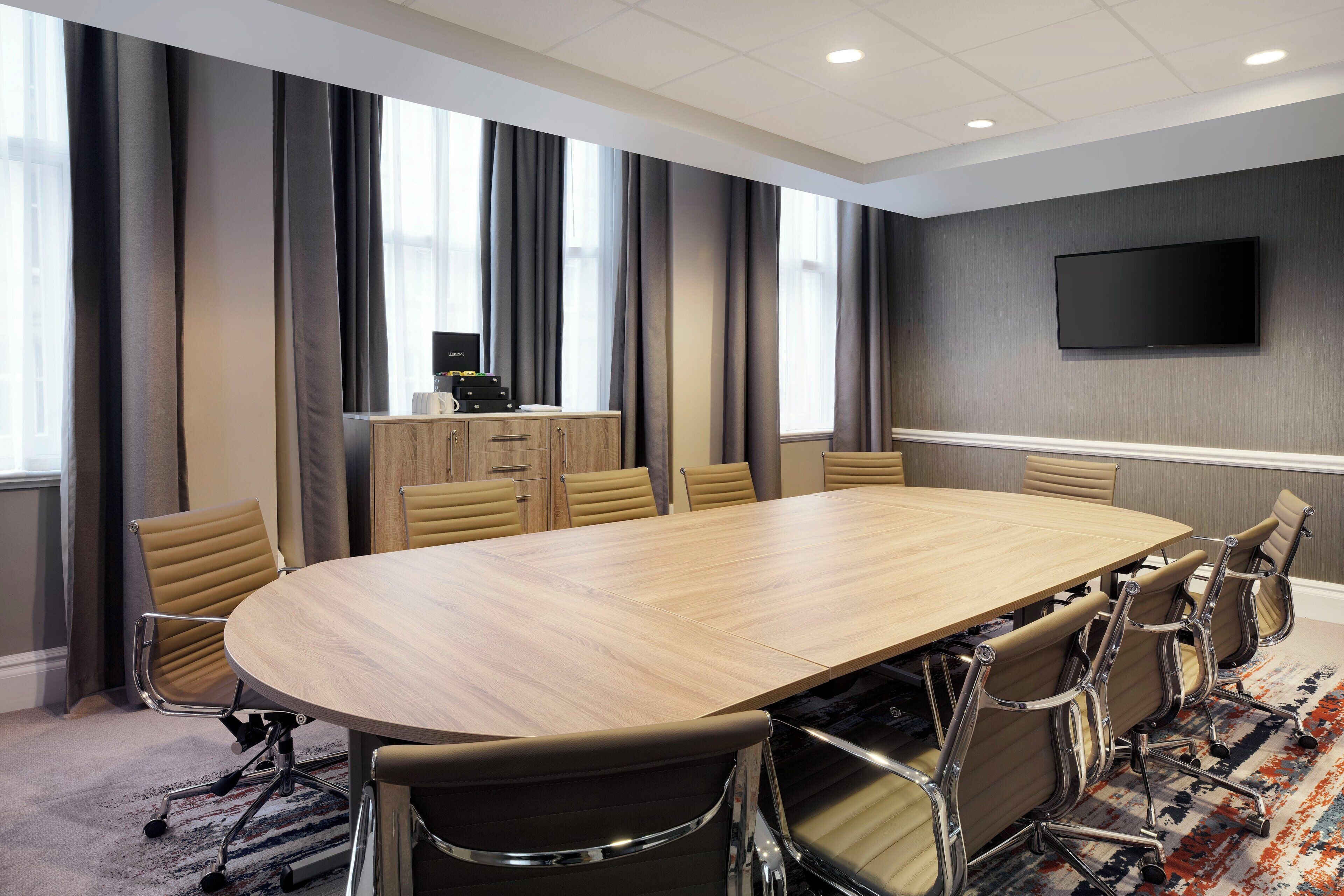 A conference room with a large oval table, six chairs, a flat-screen TV on the wall, and a cabinet with a computer and other items.