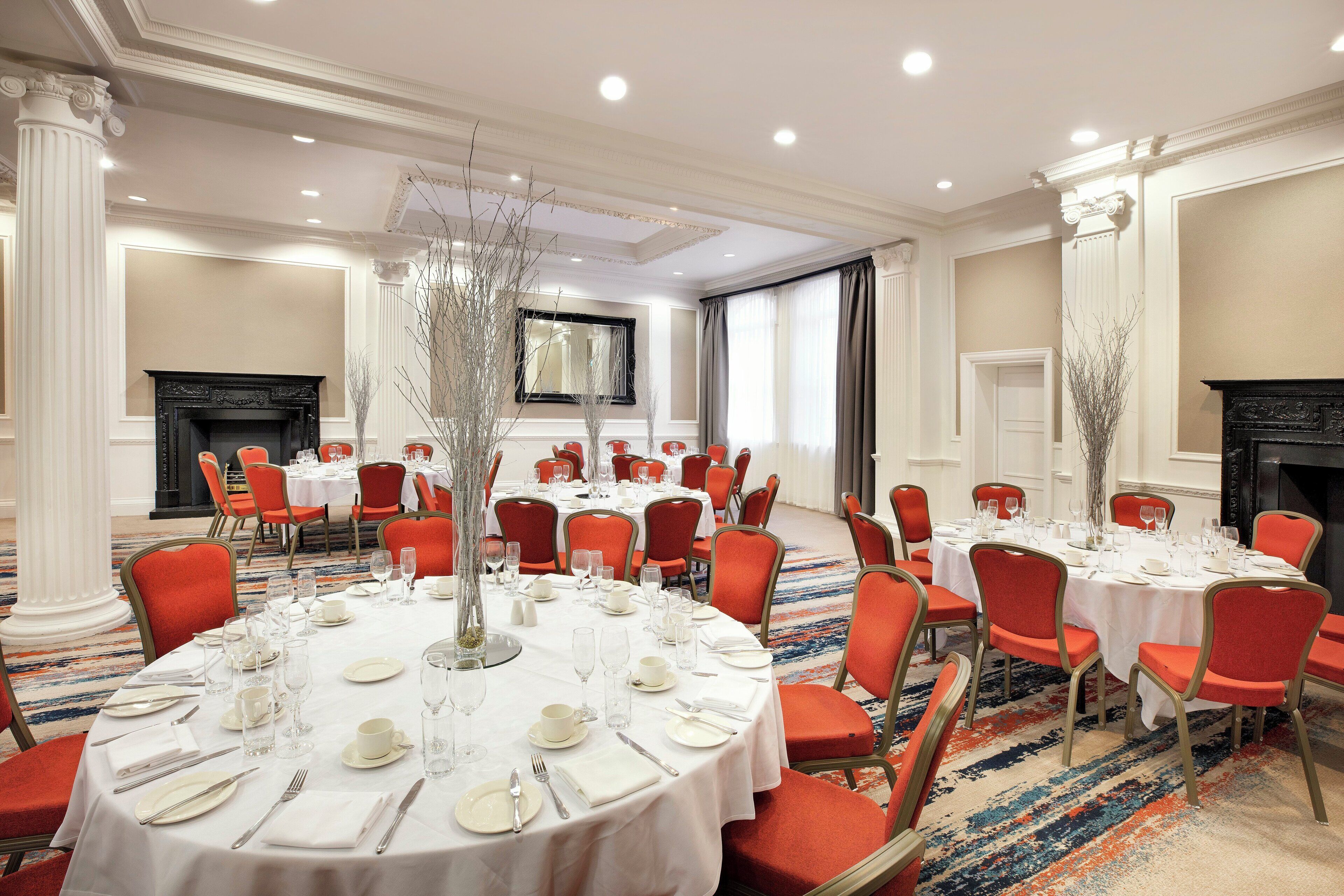 Uma sala de banquetes com mesas redondas preparadas para refeições, cadeiras vermelhas e uma lareira.