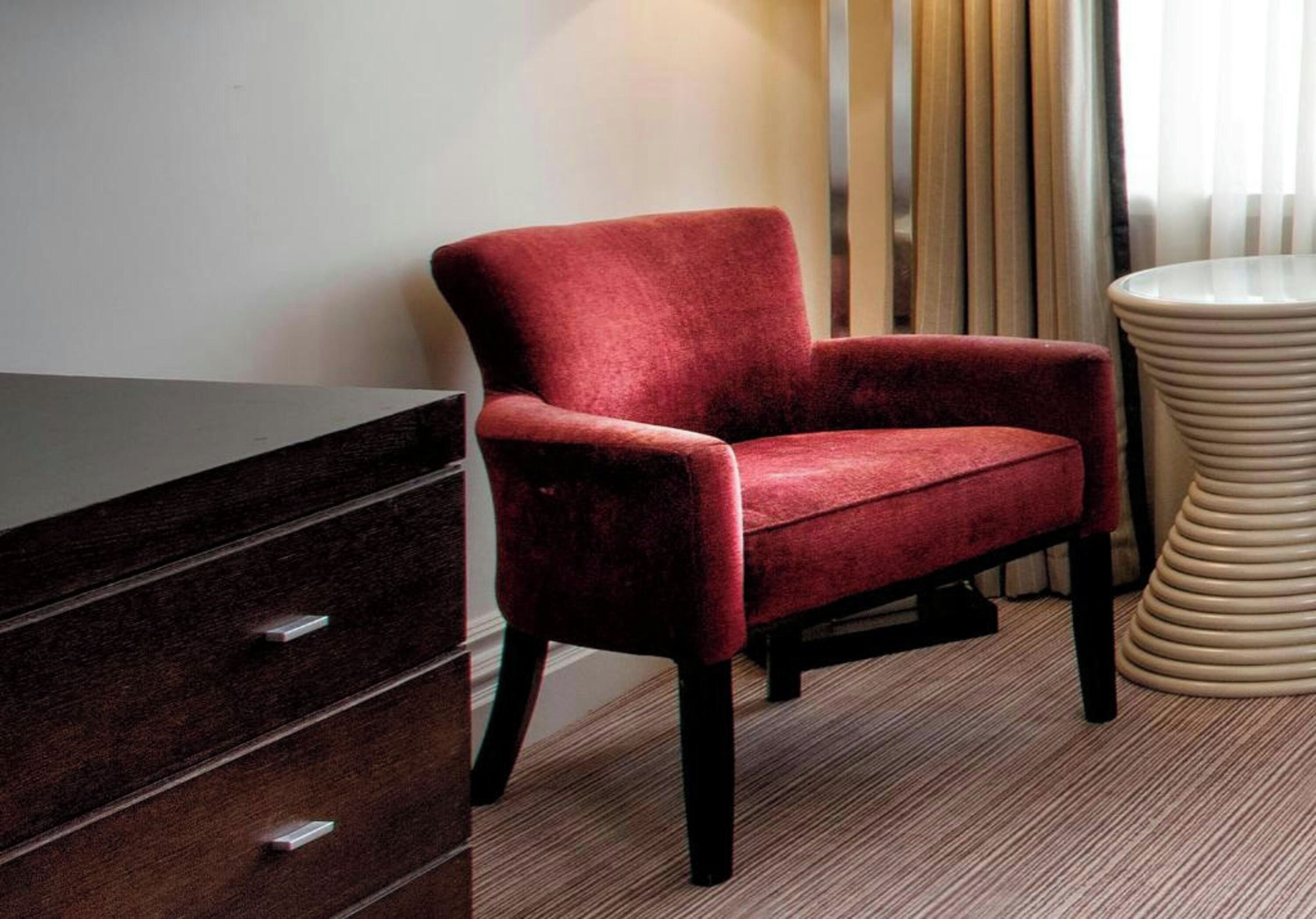 A red armchair next to a dark wooden dresser and a beige round side table in a room with striped carpet.