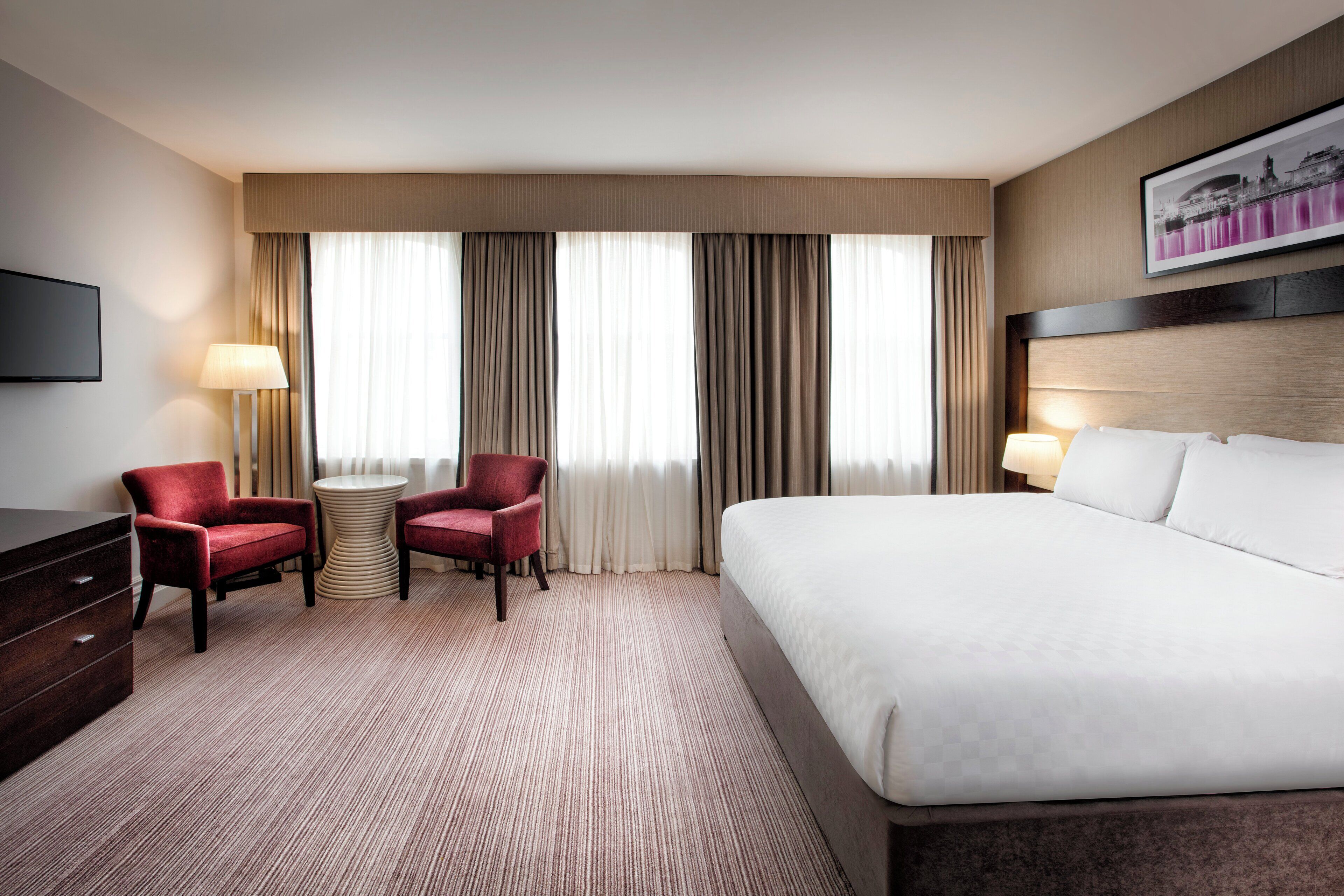Ein Hotelzimmer mit einem großen Bett, zwei Sesseln, einem kleinen Tisch, einem Fernseher und einem Gemälde an der Wand.