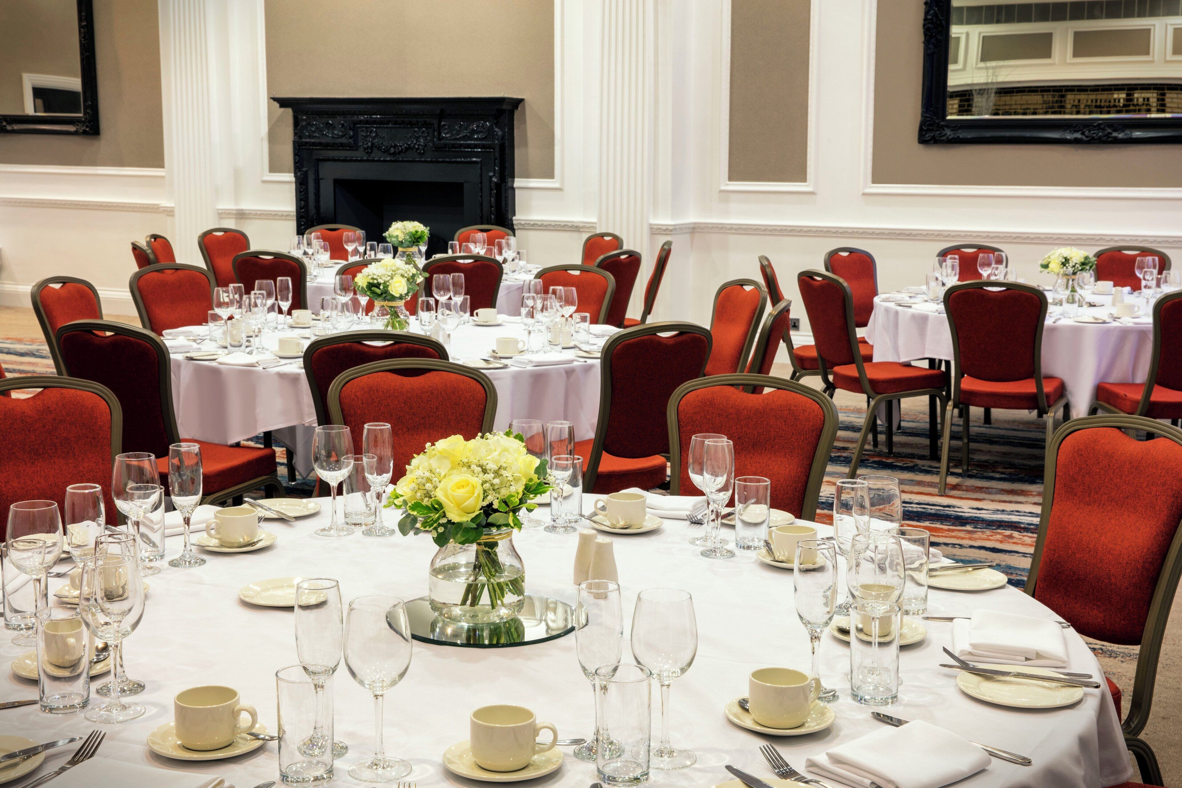 A banquet hall with round tables set for dining, red upholstered chairs, and a fireplace in the background.