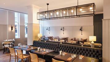 A restaurant with a high ceiling, black upholstered booths, wooden tables, and wicker chairs.