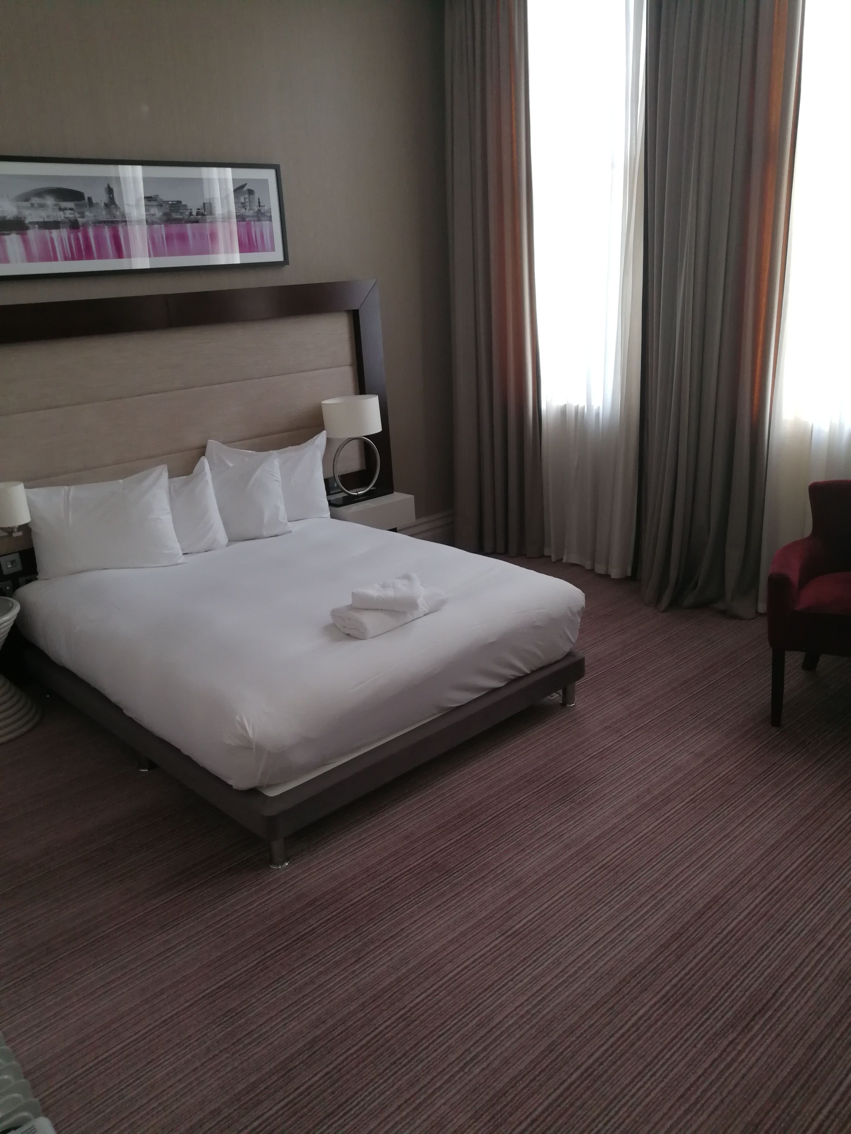A hotel room with a large bed, a headboard, a nightstand with a lamp, and a red chair.