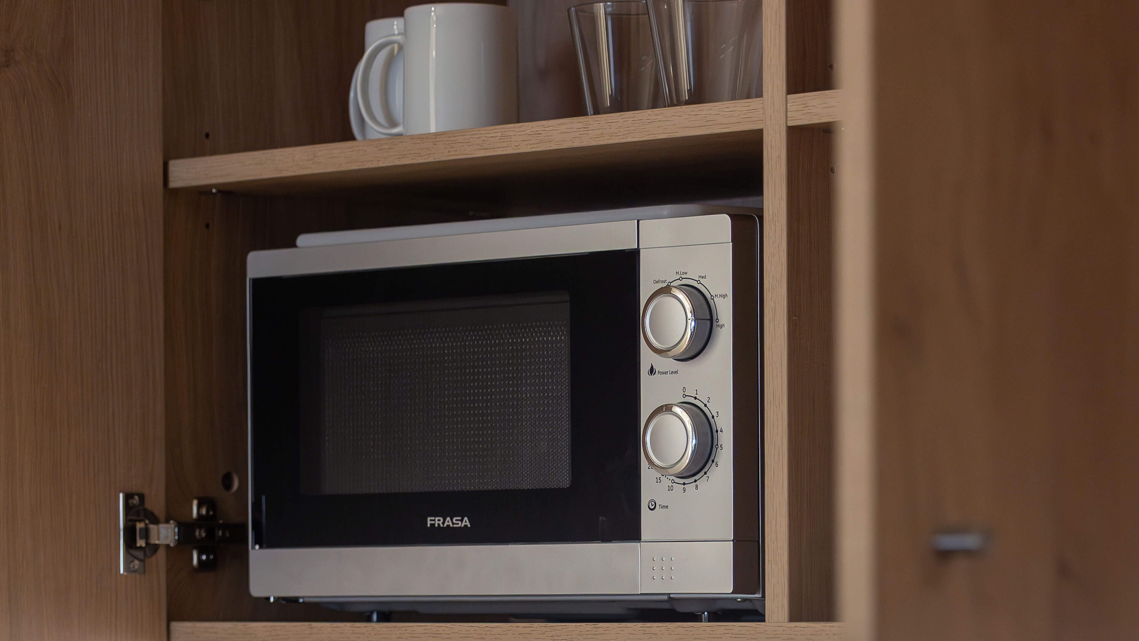 A FRASA microwave in a wooden cabinet with a shelf above containing a white mug and a clear glass.