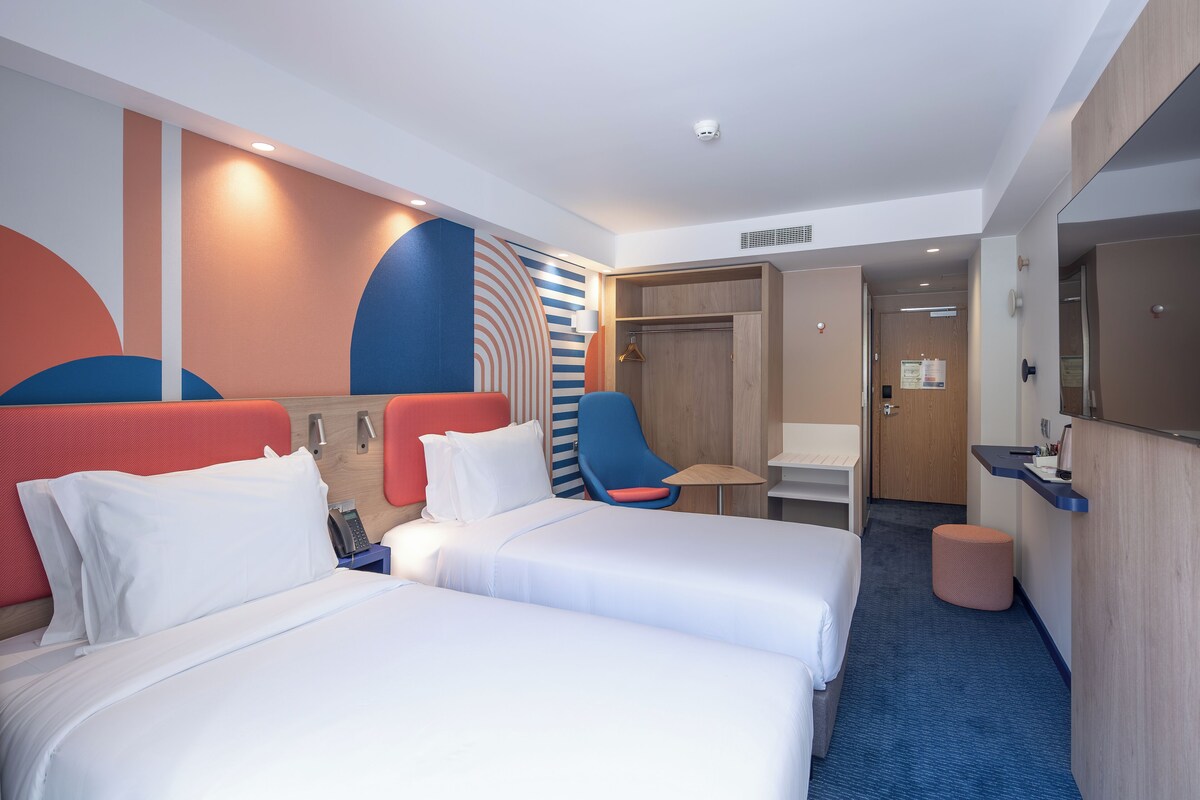 Habitación de hotel con dos camas, un escritorio, una silla, un armario y un taburete pequeño.