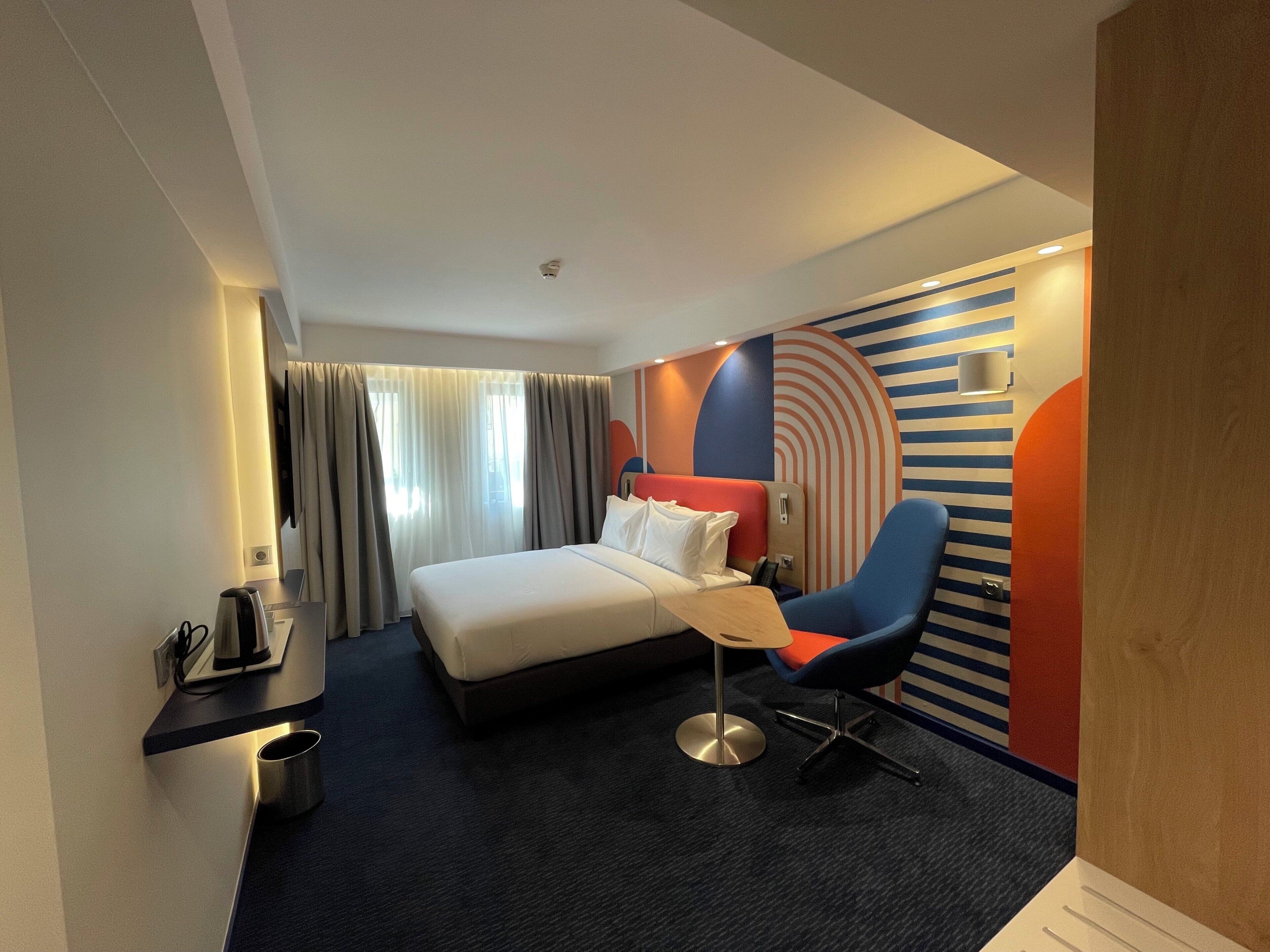 A hotel room with a bed, a chair, a desk with a phone, and a wall with a circular design.