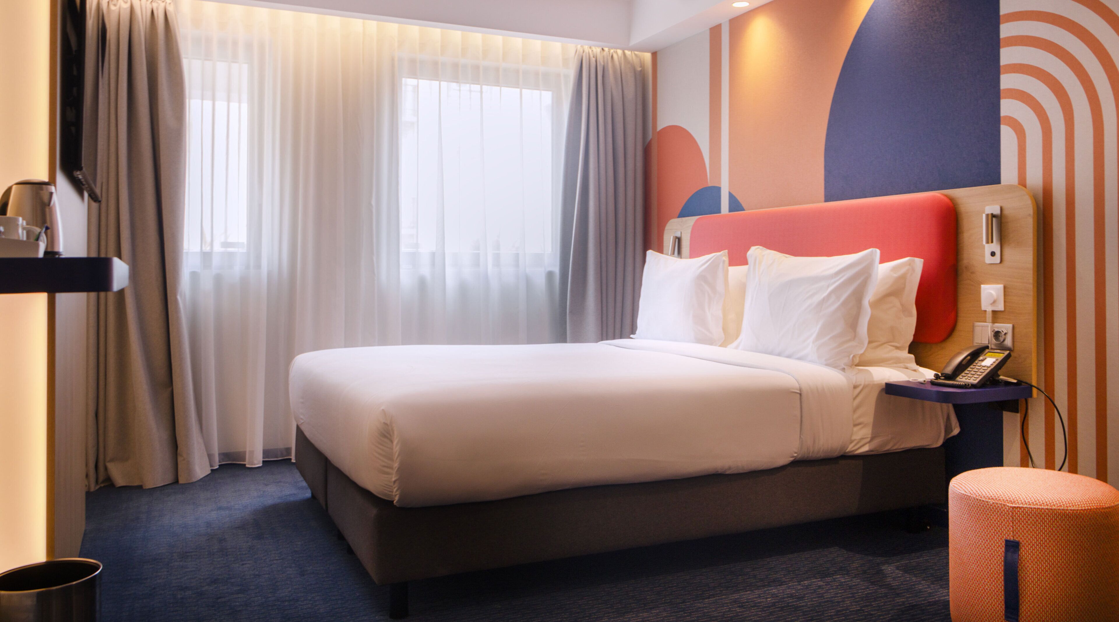 Een hotelkamer met een bed, gordijnen, een nachtkastje, een telefoon en een muur met abstract ontwerp.