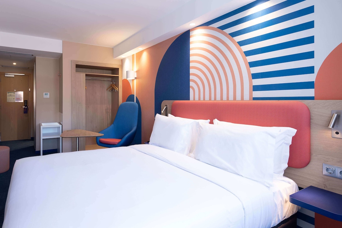 Habitación de hotel con cama, escritorio, silla y pared con diseño geométrico.