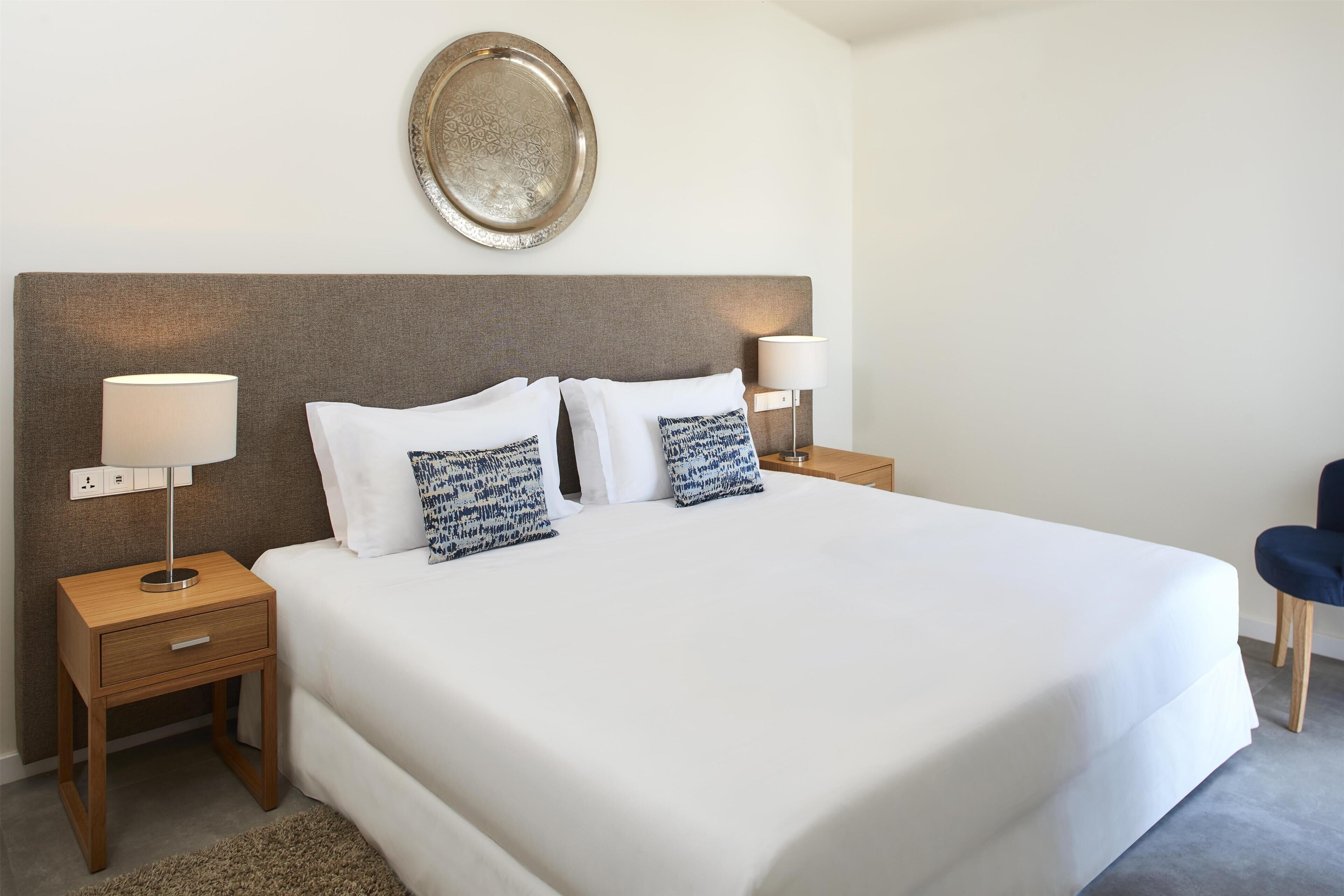 Una cama bien tendida con sábanas blancas y almohadones estampados, dos lámparas de noche, un adorno mural y una mesita de noche de madera con un cajón.