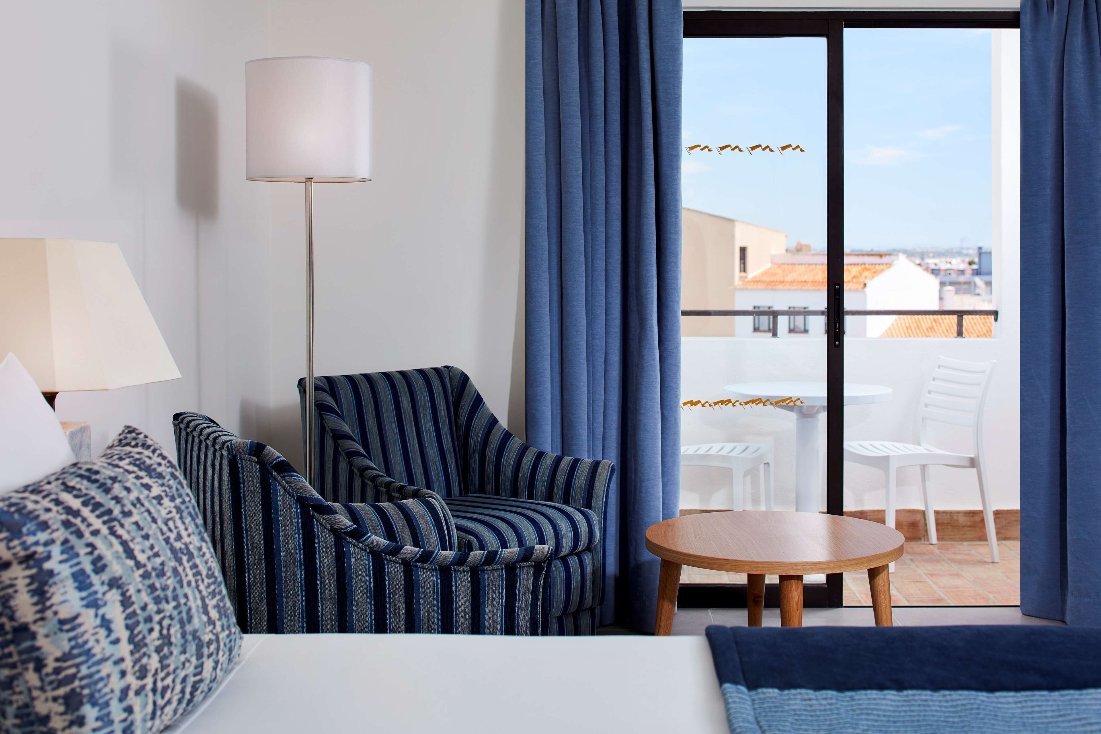 A hotel room with a bed, a striped armchair, a small table, and a view of the city through a glass door.