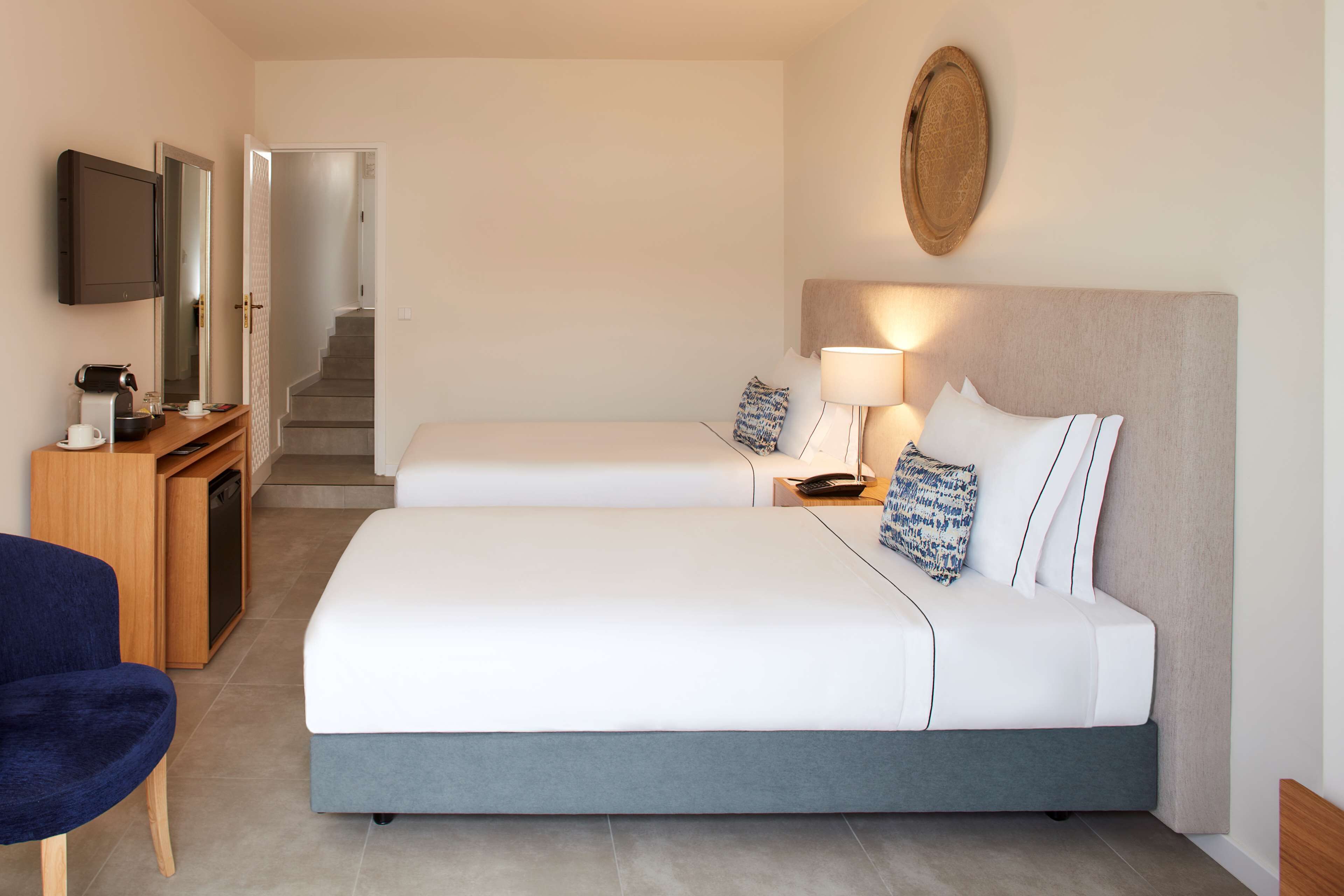 A hotel room with a bed, bedside tables, a desk with a coffee maker, a chair, and a television.