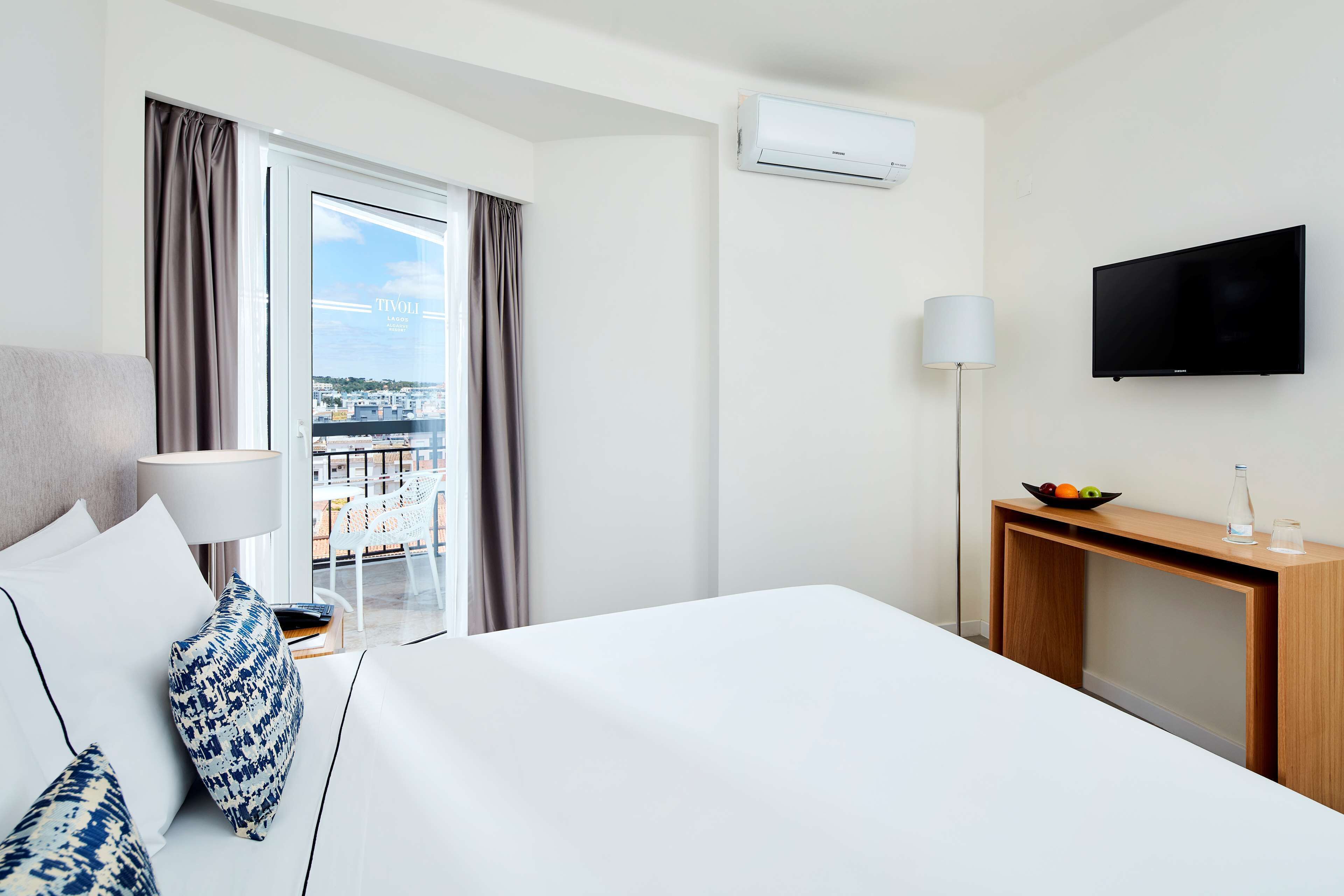 A hotel room with a bed, a desk, a TV, an air conditioner, a lamp, and a view of the city.