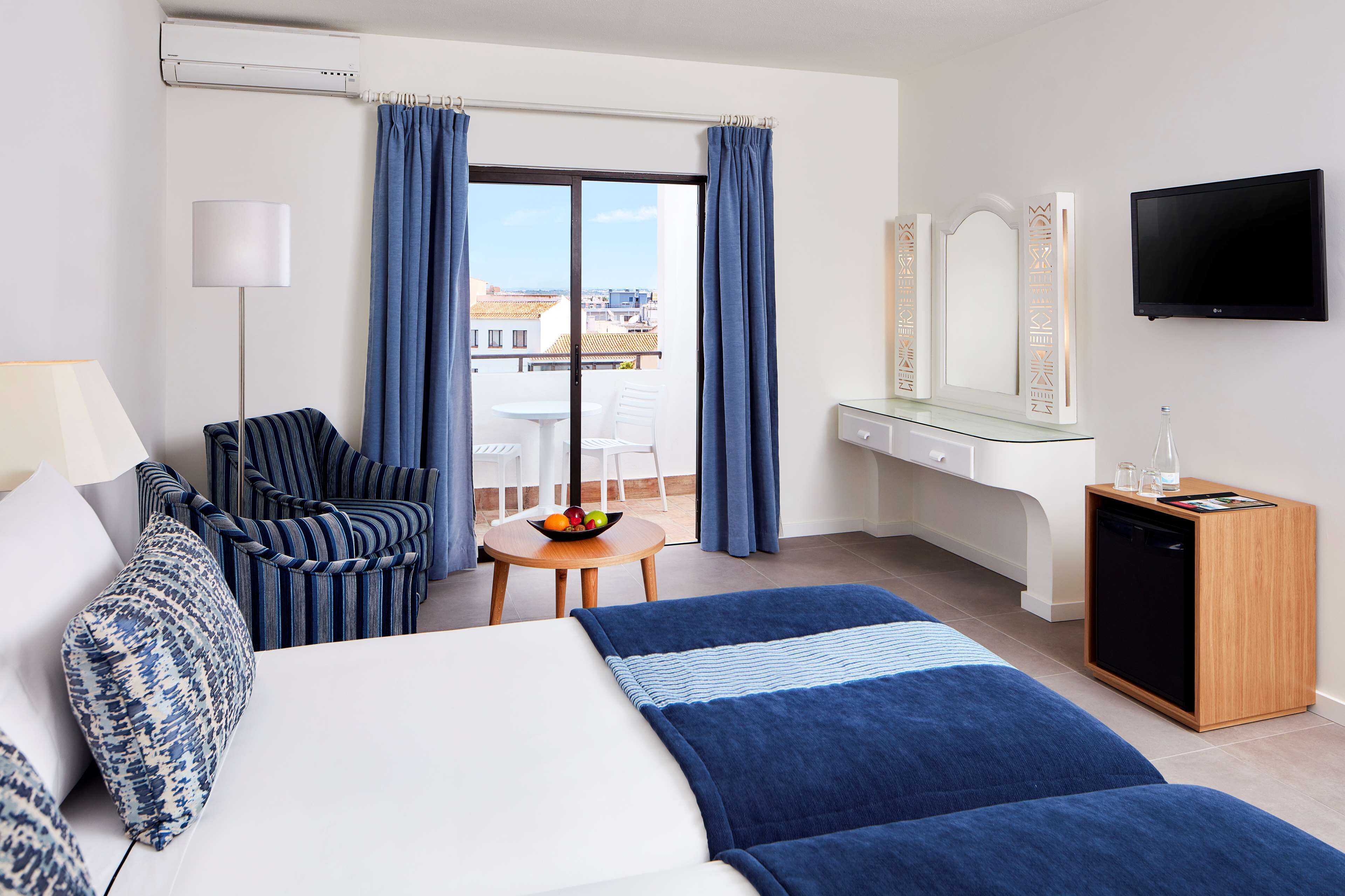A hotel room with a bed, a desk, a chair, a TV, a small table with fruit, and a balcony with curtains.