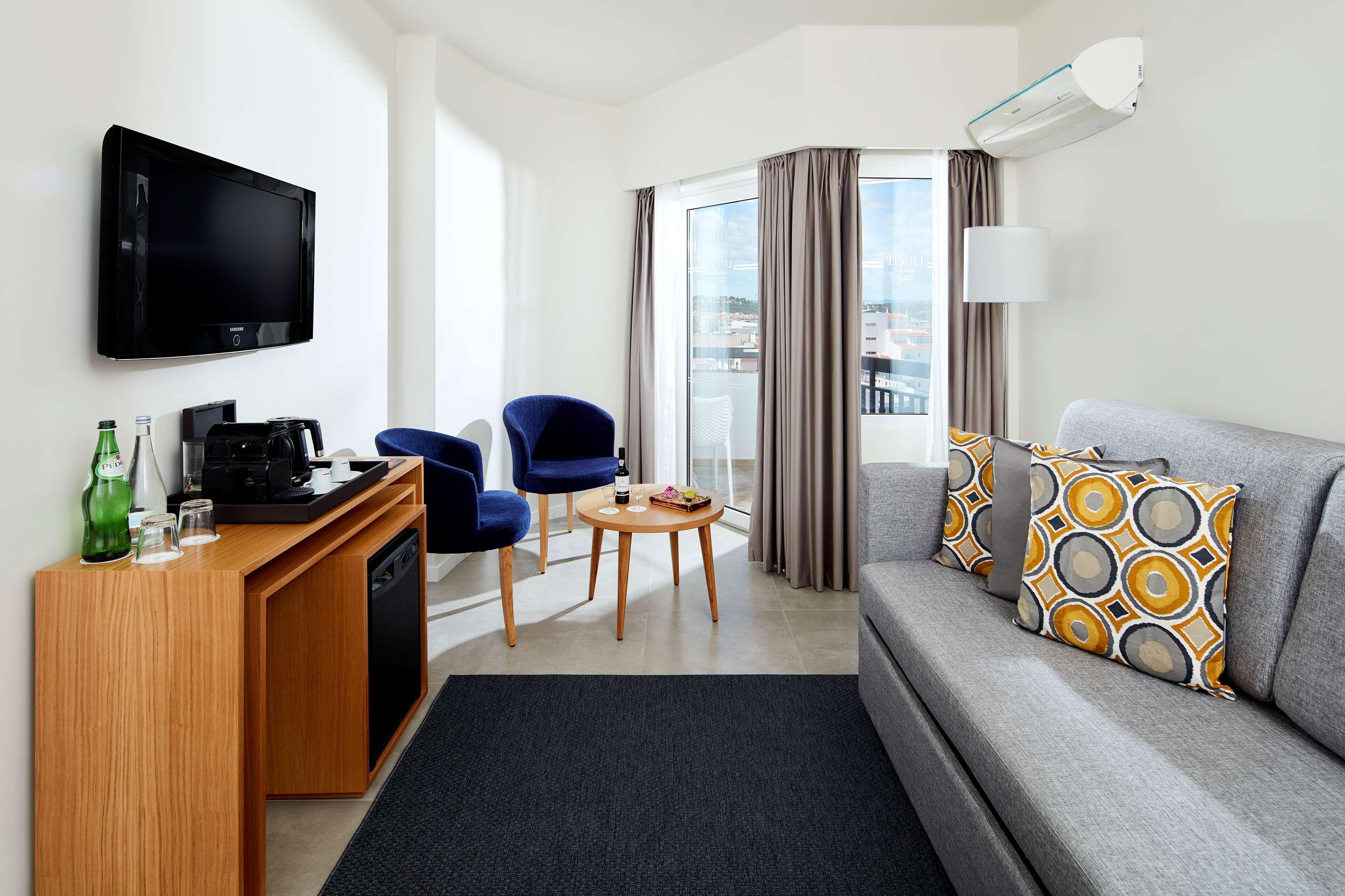 Habitación de hotel moderna con televisión de pantalla plana, sof á con cojines estampados, estación de café, mesa de comedor y vistas a través de un ventanal grande.
