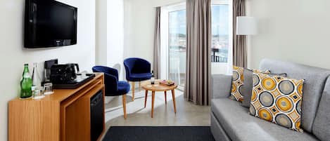 Une chambre d’hôtel moderne dotée d’une télévision à écran plat, d’un canapé avec des coussins à motifs, d’un coin café, d’une table à manger et offrant une vue sur l’extérieur grâce à une grande fenêtre.