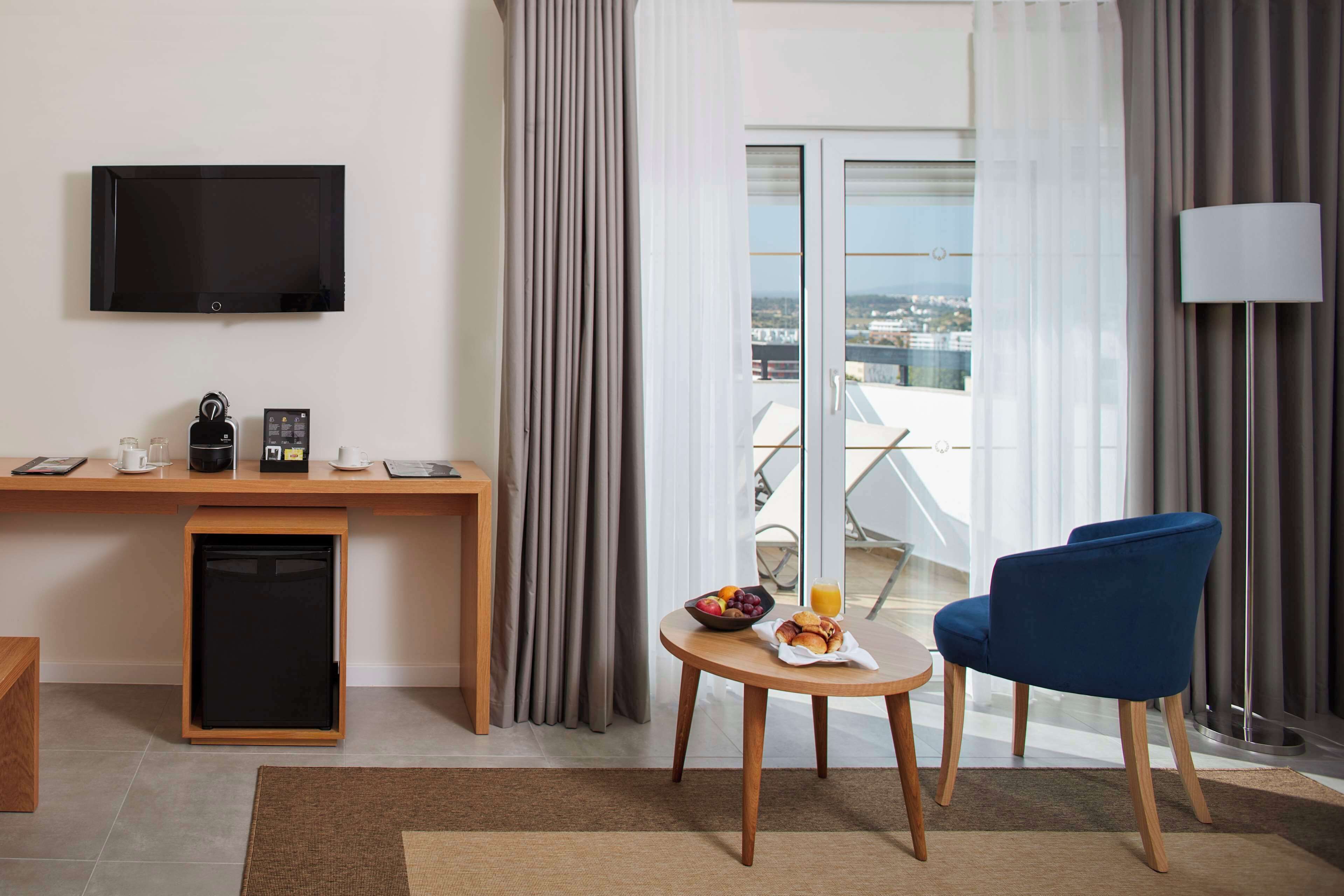 A hotel room with a flat-screen TV, a small refrigerator, a wooden desk, a blue chair, a round table with fruits and a coffee cup, and a view of the city through large windows.