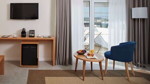 A hotel room with a flat-screen TV, a small refrigerator, a wooden desk, a blue chair, a round table with fruits and a coffee cup, and a view of the city through large windows.