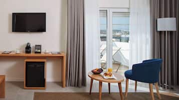 A hotel room with a flat-screen TV, a small refrigerator, a wooden desk, a blue chair, a round table with fruits and a coffee cup, and a view of the city through large windows.