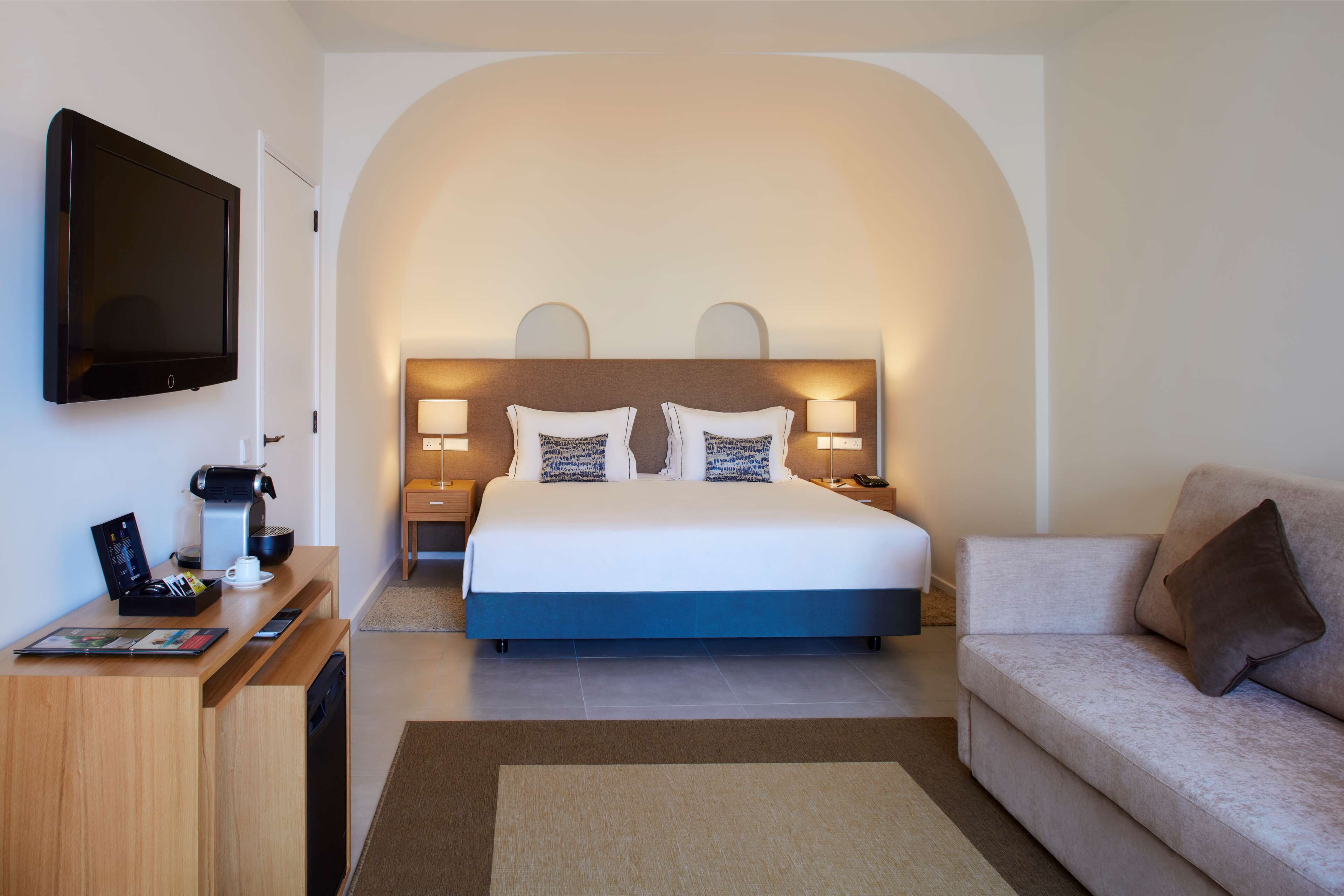 A hotel room with a bed, a TV, a desk with a coffee maker, a sofa, and a large arched wall.