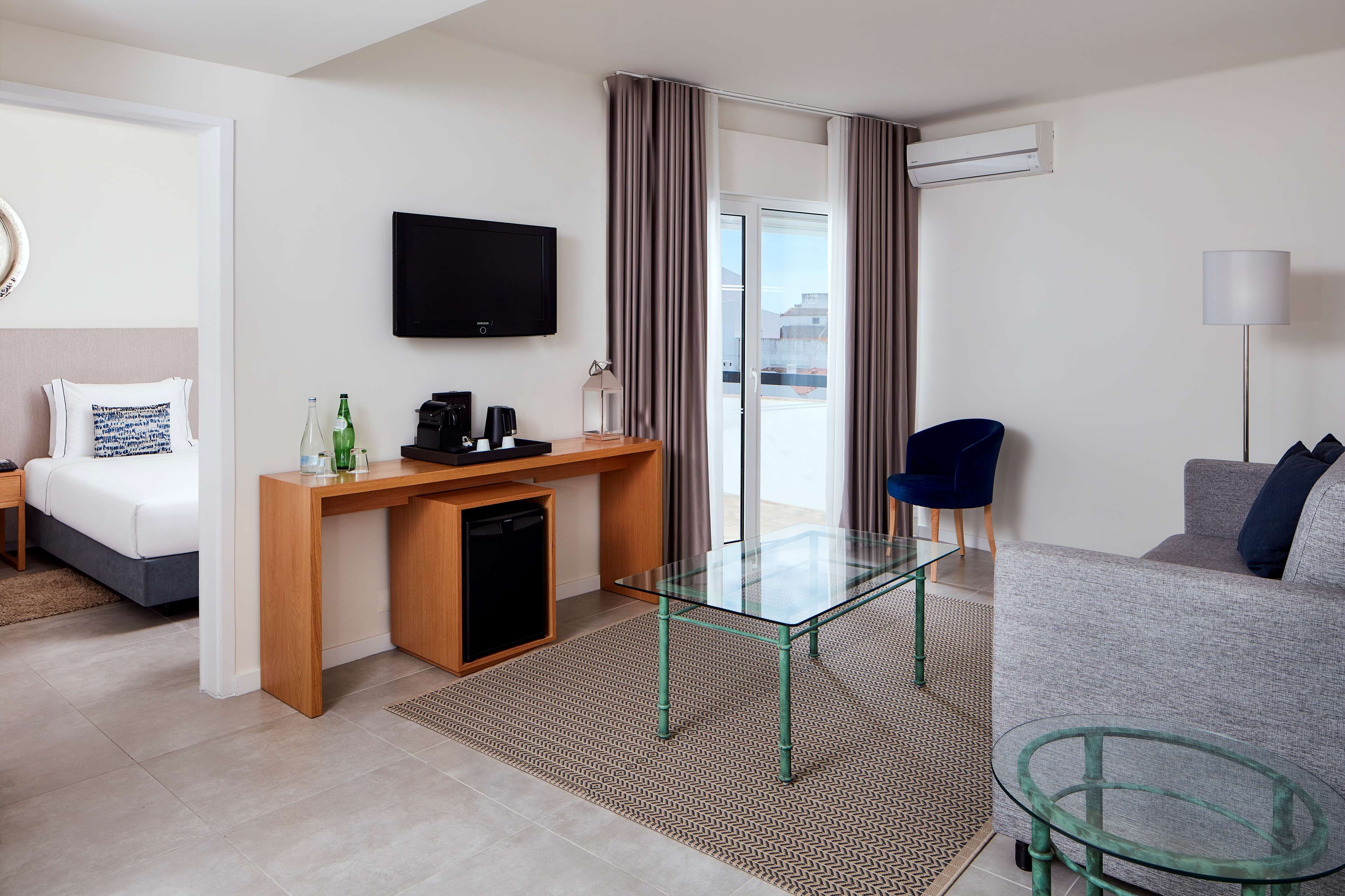 A hotel room with a bed, a desk with a television, a glass table, a sofa, and a chair.