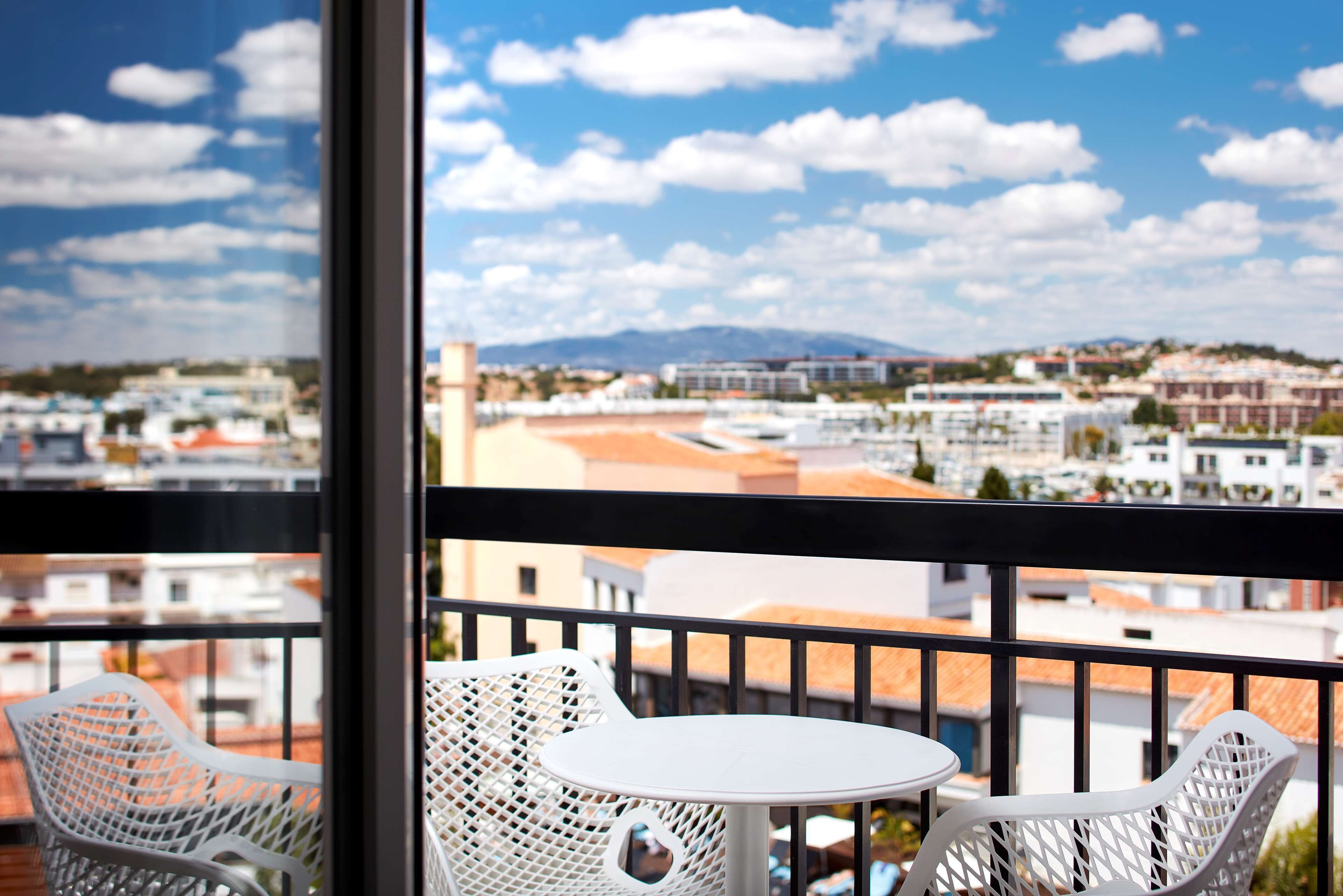 A balcony with white wicker chairs and a small round table, overlooking a cityscape with buildings and a mountain in the distance.
