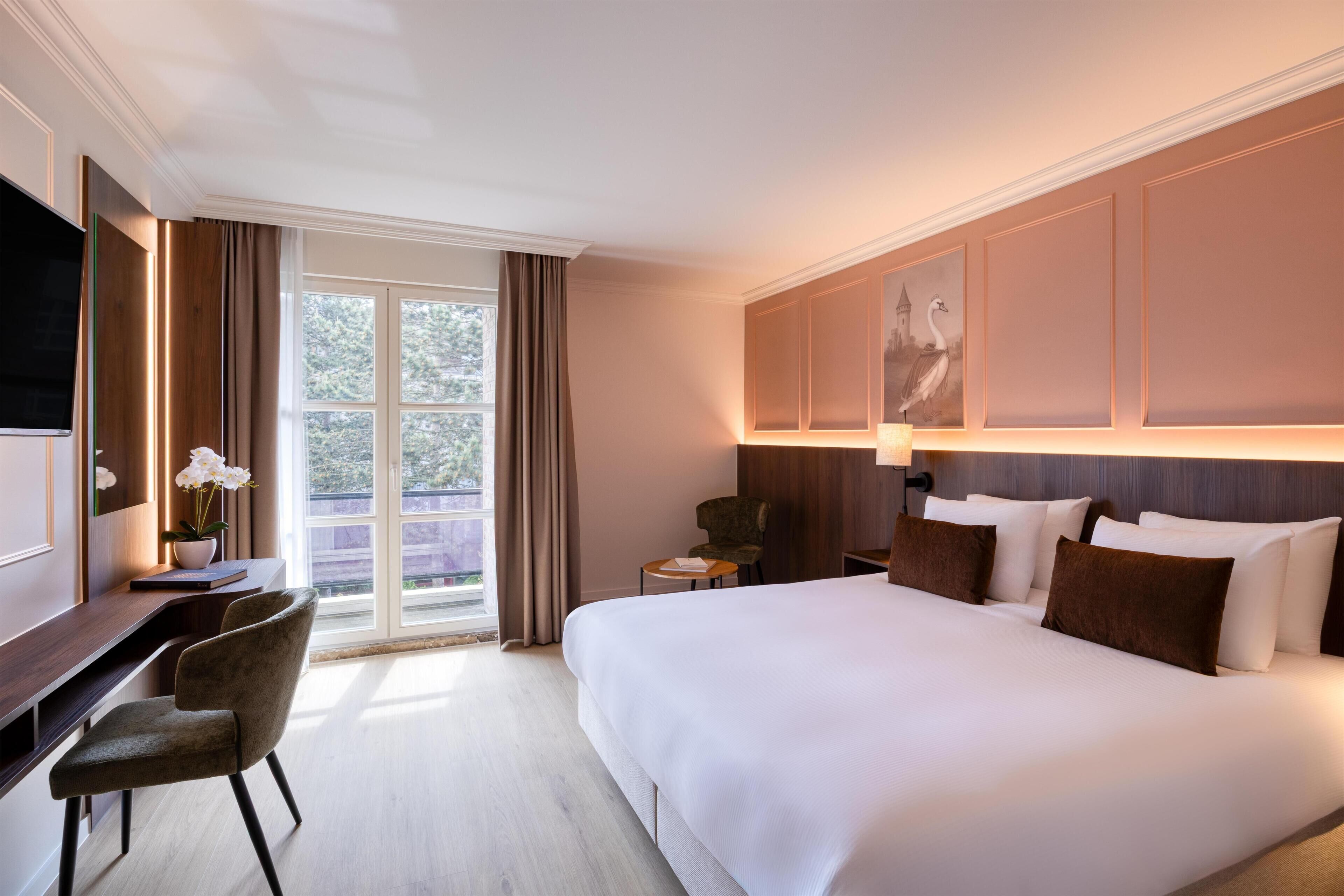 Habitación de hotel con una cama grande, un escritorio con una silla, un televisor y una ventana con cortinas.